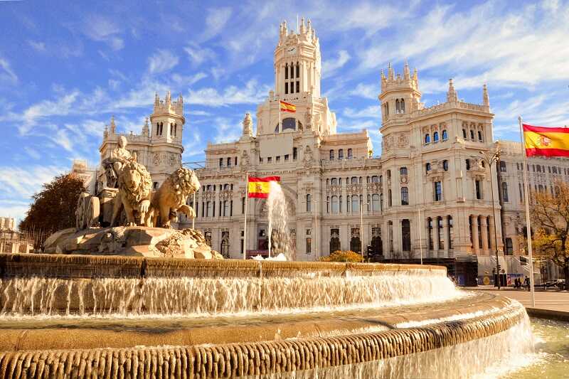 Brunnen mit Löwenstatue und dem Palacio de Cibeles im Hintergrund, umgeben von spanischen Flaggen und blauem Himmel.