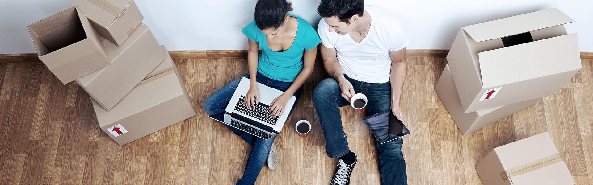 Zwei Personen sitzen auf dem Boden mit Umzugskartons und einem Laptop, während sie Kaffee trinken.
