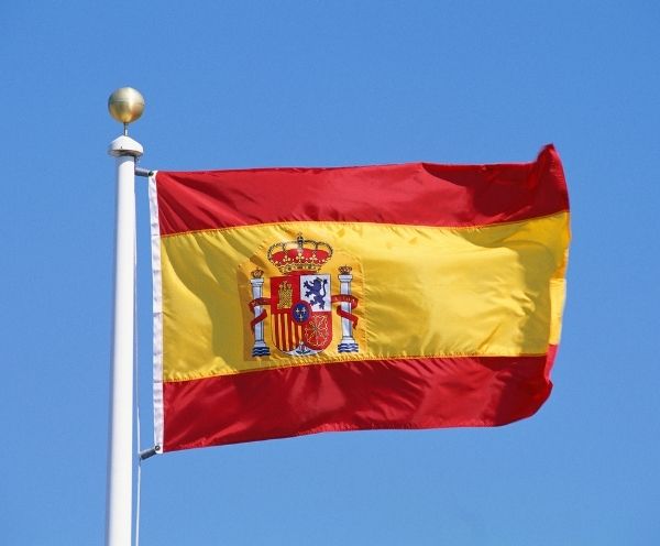 Flagge Spaniens mit rot-gelbem Design und Wappen auf einem Fahnenmast vor klarem Himmel.