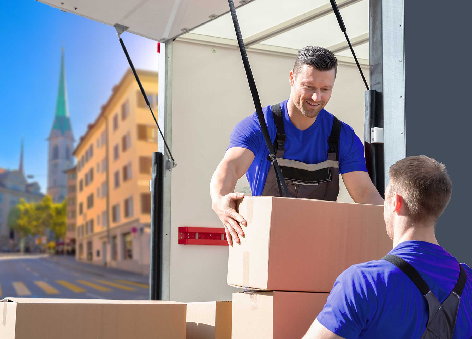 Mann in blauer T-Shirt und Latzhose hebt Kartons aus einem Umzugswagen in einer Stadt mit einem Kirchturm im Hintergrund.